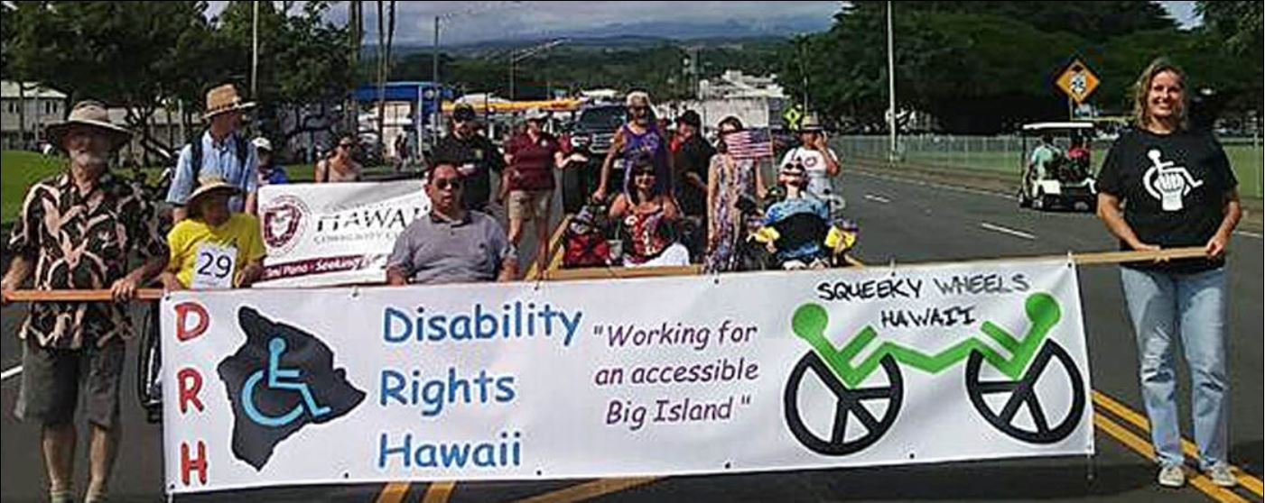 A picture of about 15 DRH members marching in the November 10, 2018, Veterans Day Parade in Hilo. A banner is being carried at the front of the marchers announcing Disability Rights Hawaii (DRH) and the Facebook group named Squeaky Wheels Hawaii. Logos representing the two groups are printed on the banner (an outline map of Hawaii County with a wheelchair symbol inside it) and Squeaky Wheels (two wheelchairs facing each other, each performing a wheelie). Many of the members are marching with wheelchairs.
